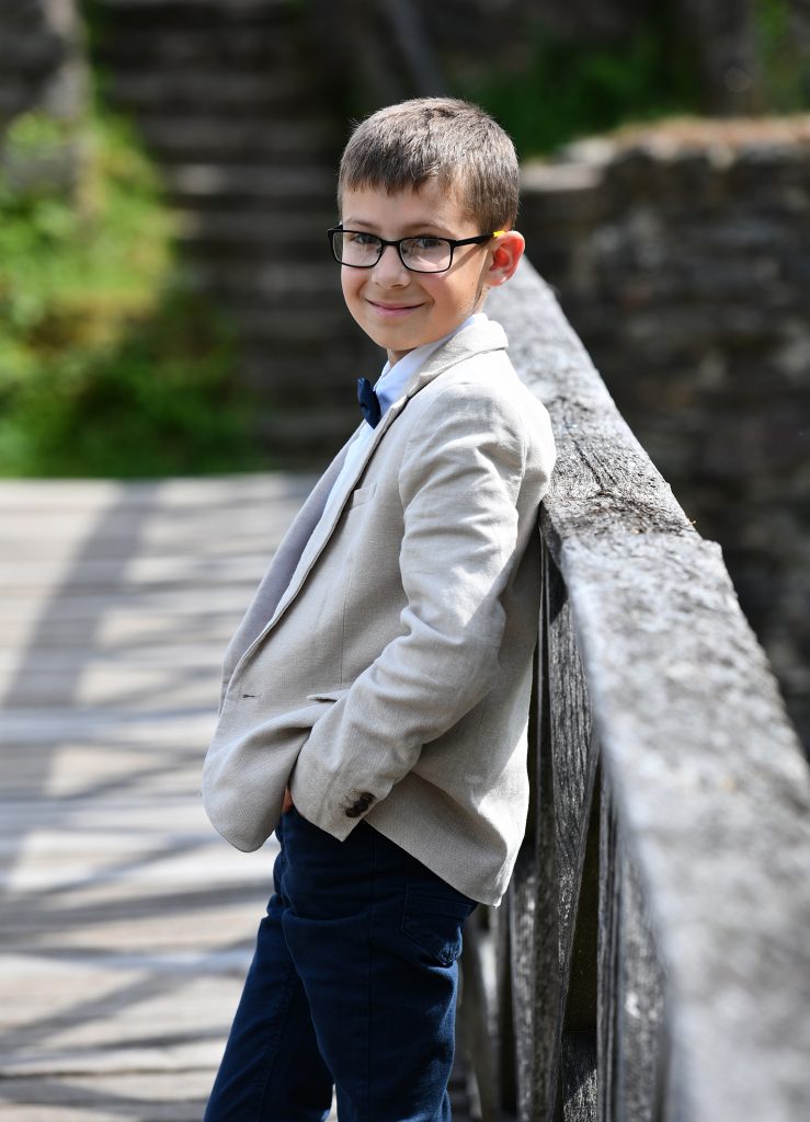 Natürliche Kommunionfotos - Junge in heller Anzugjacke lehnt lässig an einer Holzgeländer