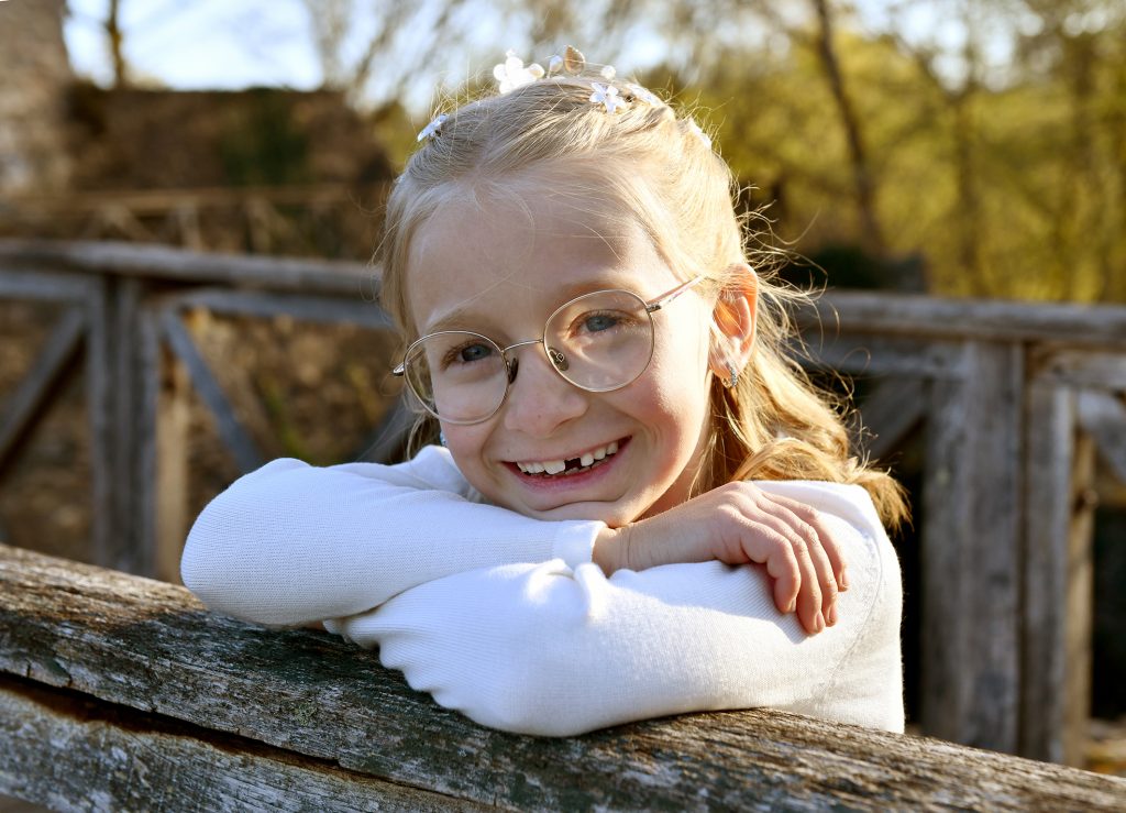 Natürliche Kommunionfotos - lachendes Mädchen in weißen Kommunionkleid auf einem Holz-Brückengeländer